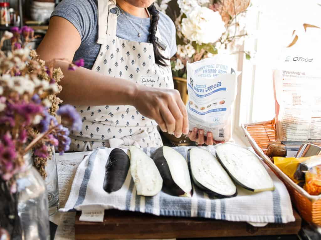 A person in a gray shirt and patterned apron is seasoning sliced eggplants on a towel-covered board. Surrounding the scene are flowers and cooking items, with sunlight streaming in from the side.